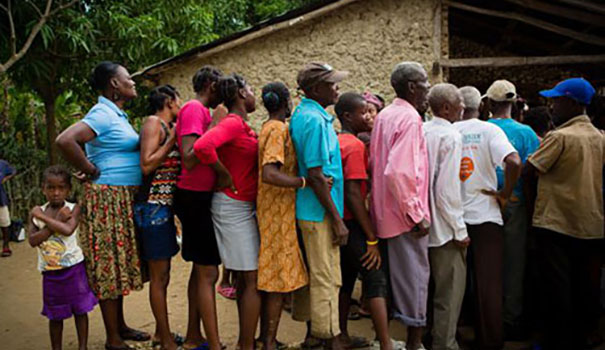 People standing in a cue outside a stone and wooden building in a jungle.